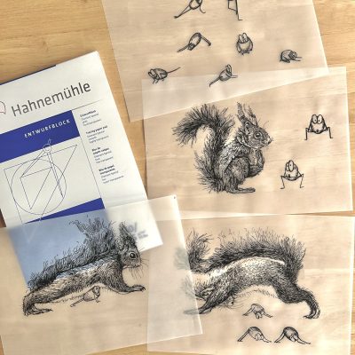 Vier Blätter Transparentpapier mit Tintezeichnungen liegen übereinander auf einem Holztisch. Links oben ein Hahnemühle Entwurfsblock.