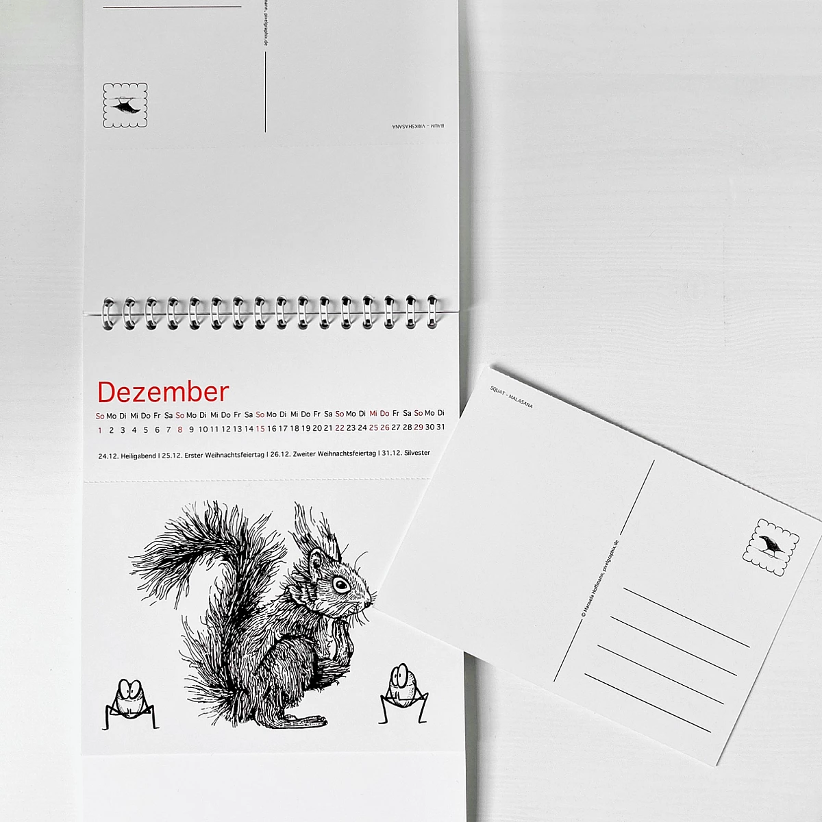 Der Kalender Ommmmmmmmmmmm - Yoga für Eichhörnchen & ähnlich veranlagte Menschen - Postkartenkalender 2024  steht seitlich aufgeklappt auf einer weißen Fläche. Auf dem Cover ist ein Eichhörnchen zu sehen, das die Position des herabschauenden Hundes praktiziert.  Fünf Haselnüsse praktizieren ebenfalls Yoga.