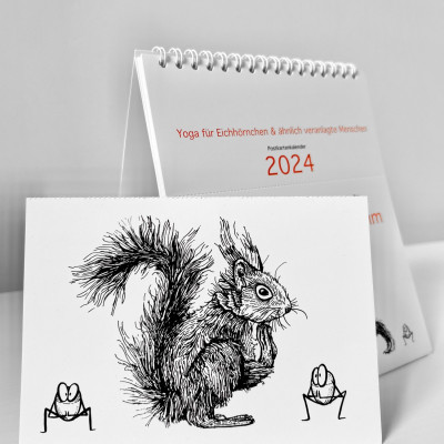 Der Kalender „Ommmmmmmmmmmm - Yoga für Eichhörnchen & ähnlich veranlagte Menschen - Postkartenkalender 2024" steht aufgeklappt auf einer weißen Fläche. Davor steht eine abgetrennte Postkarte. Die schwarze Illustration zeigt ein Eichhörnchen und zwei Haselnüsse in Squat-Pose.