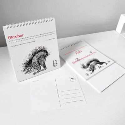 Der Kalender Ommmmmmmmmmmm, Postkartenkalender 2024 - steht aufgestellt auf einem Tisch, ein Kalender liegt ungefaltet daneben. Im Vordergrund liegt eine abgetrennte Postkarte.