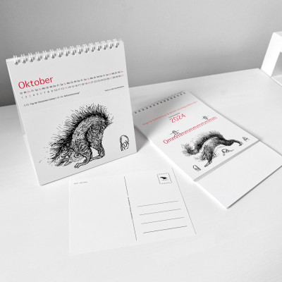Der Kalender Ommmmmmmmmmmm, Postkartenkalender 2024 - steht aufgestellt auf einem Tisch, ein Kalender liegt ungefaltet daneben. Im Vordergrund liegt eine abgetrennte Postkarte.